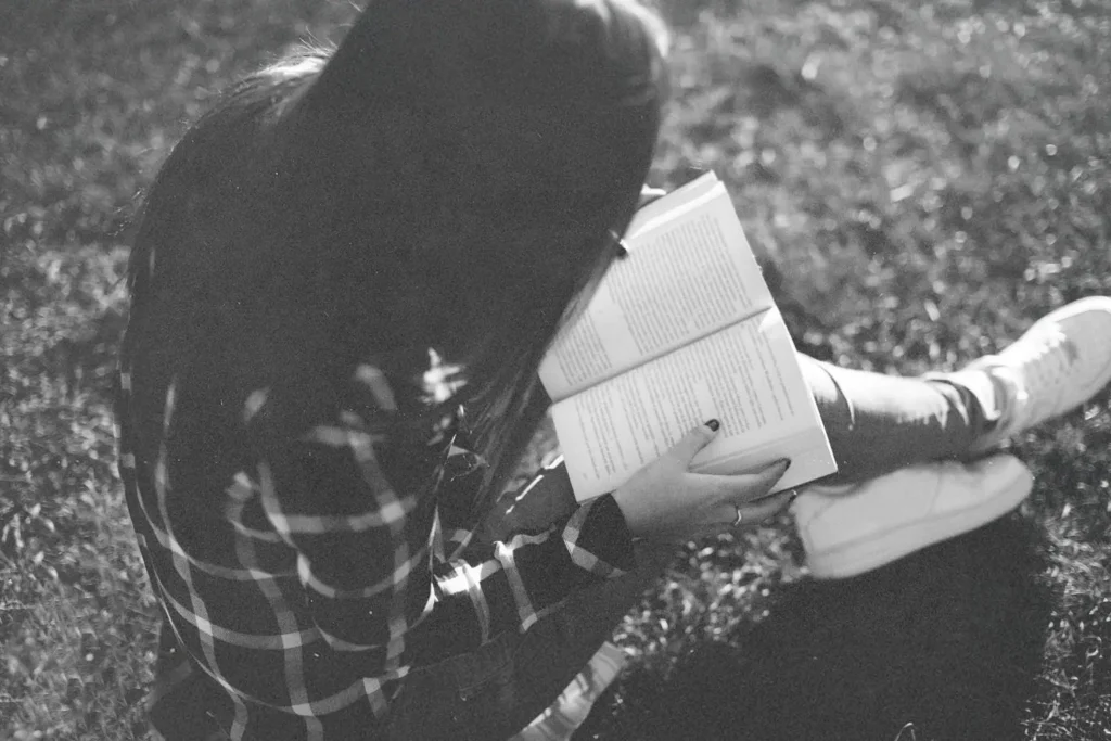 Person sitting on grass outdoors, holding an open book while reading in soft natural light