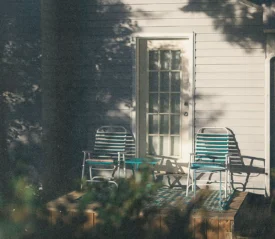 Two striped patio chairs sit on a small wooden porch outside a house, lit by warm late‑afternoon sunlight