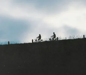 Two cyclists ride along a ridgeline, silhouetted against a cloudy sky