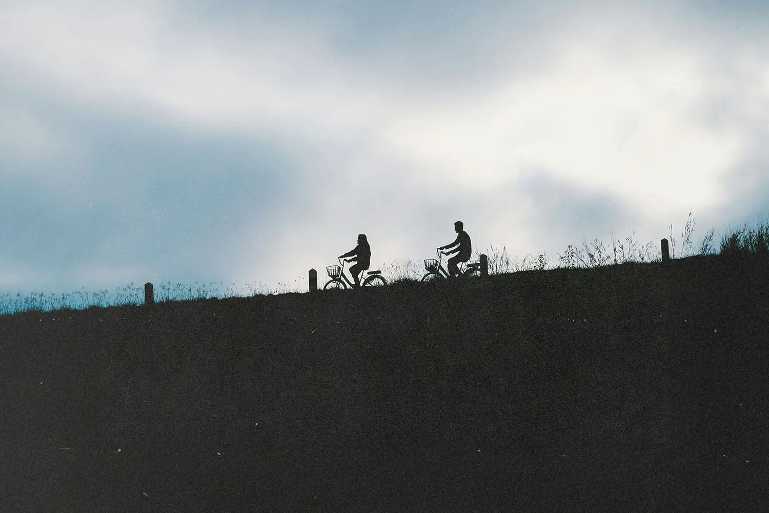 Two cyclists ride along a ridgeline, silhouetted against a cloudy sky