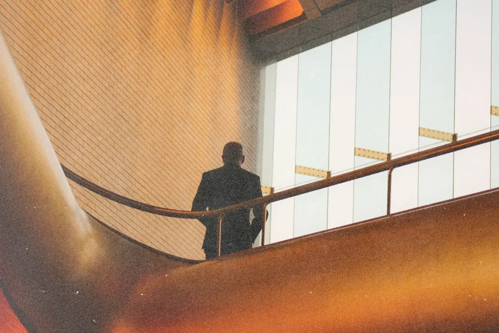 Figure leaning on a railing inside a contemporary structure with warm lighting and large windows