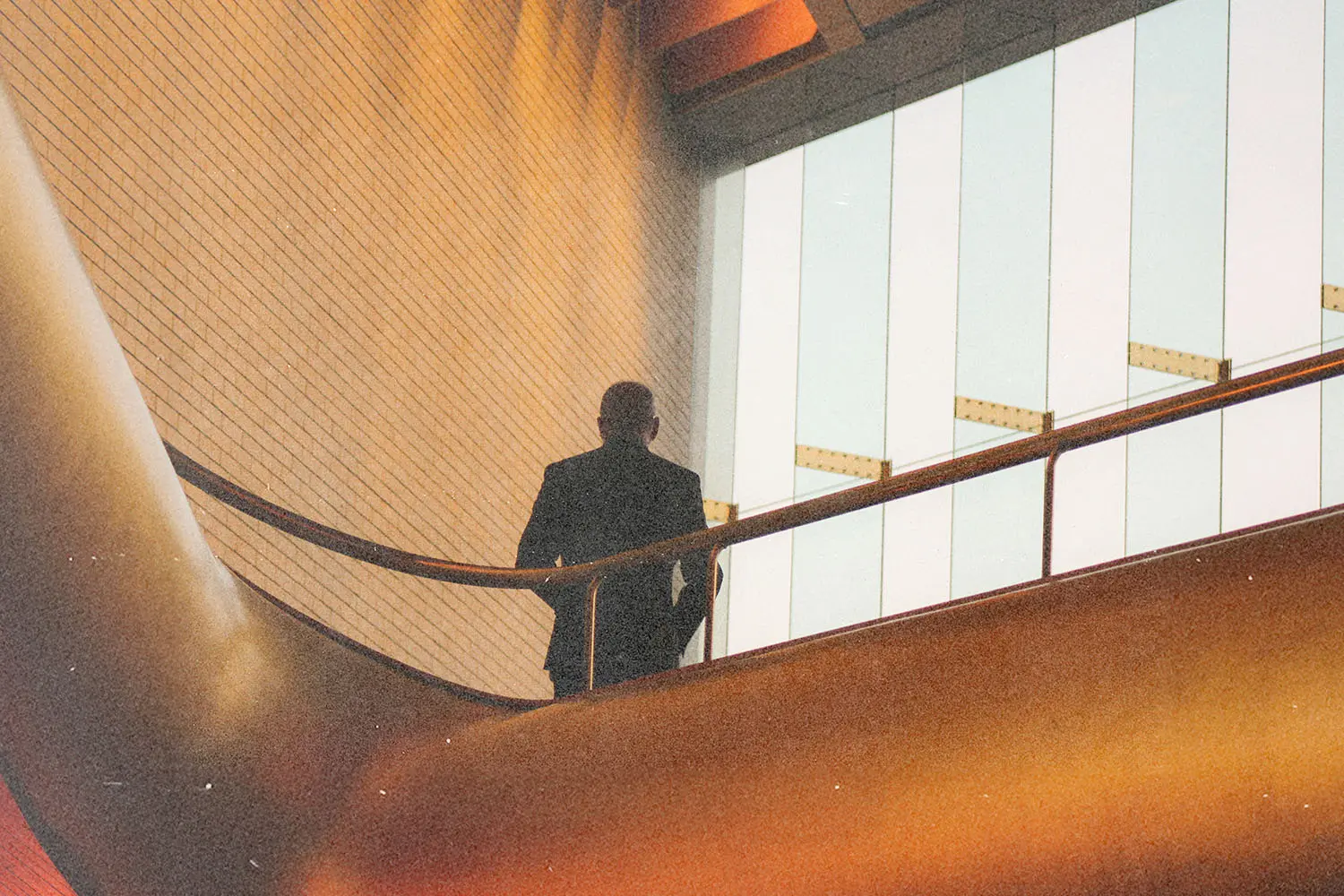 Figure leaning on a railing inside a contemporary structure with warm lighting and large windows