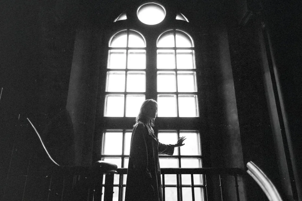 Silhouetted person standing indoors and touching a tall, bright window with arched panes, with light streaming in behind them