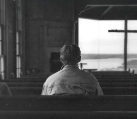 Two people sitting in church pews facing a large window with a cross silhouetted against a distant landscape, while others stand near the front