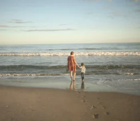 Adult and child standing side by side on the beach, looking out at the calm ocean under a pastel sky