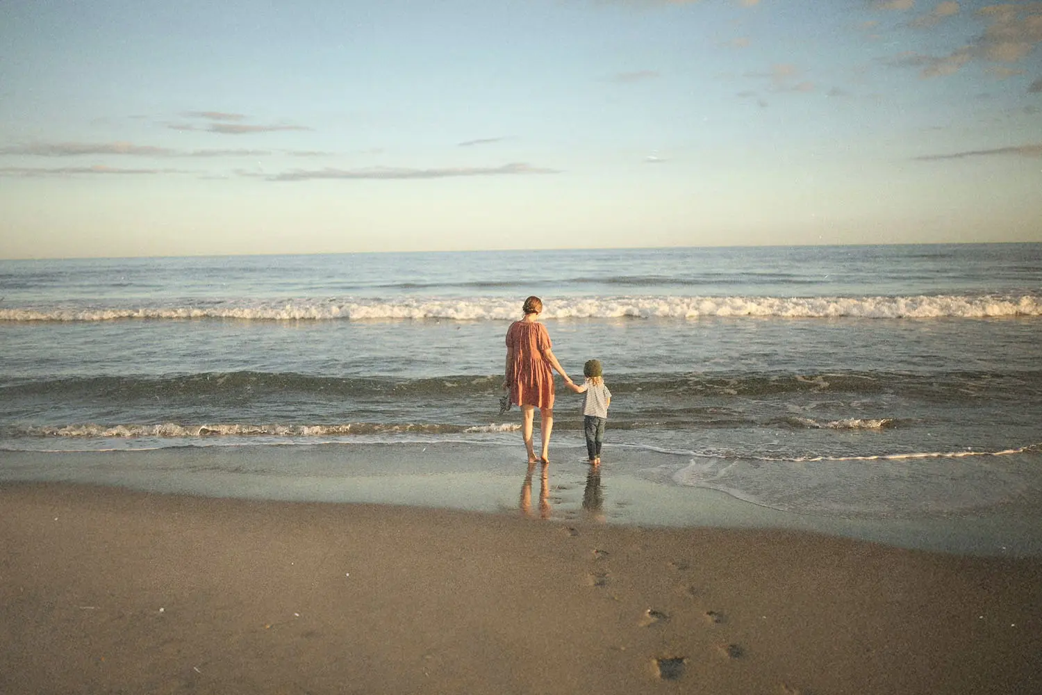 Adult and child standing side by side on the beach, looking out at the calm ocean under a pastel sky