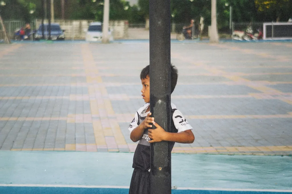 Child wearing a sports uniform stands outdoors, holding onto a tall metal pole in a paved courtyard or playground