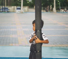Child wearing a sports uniform stands outdoors, holding onto a tall metal pole in a paved courtyard or playground