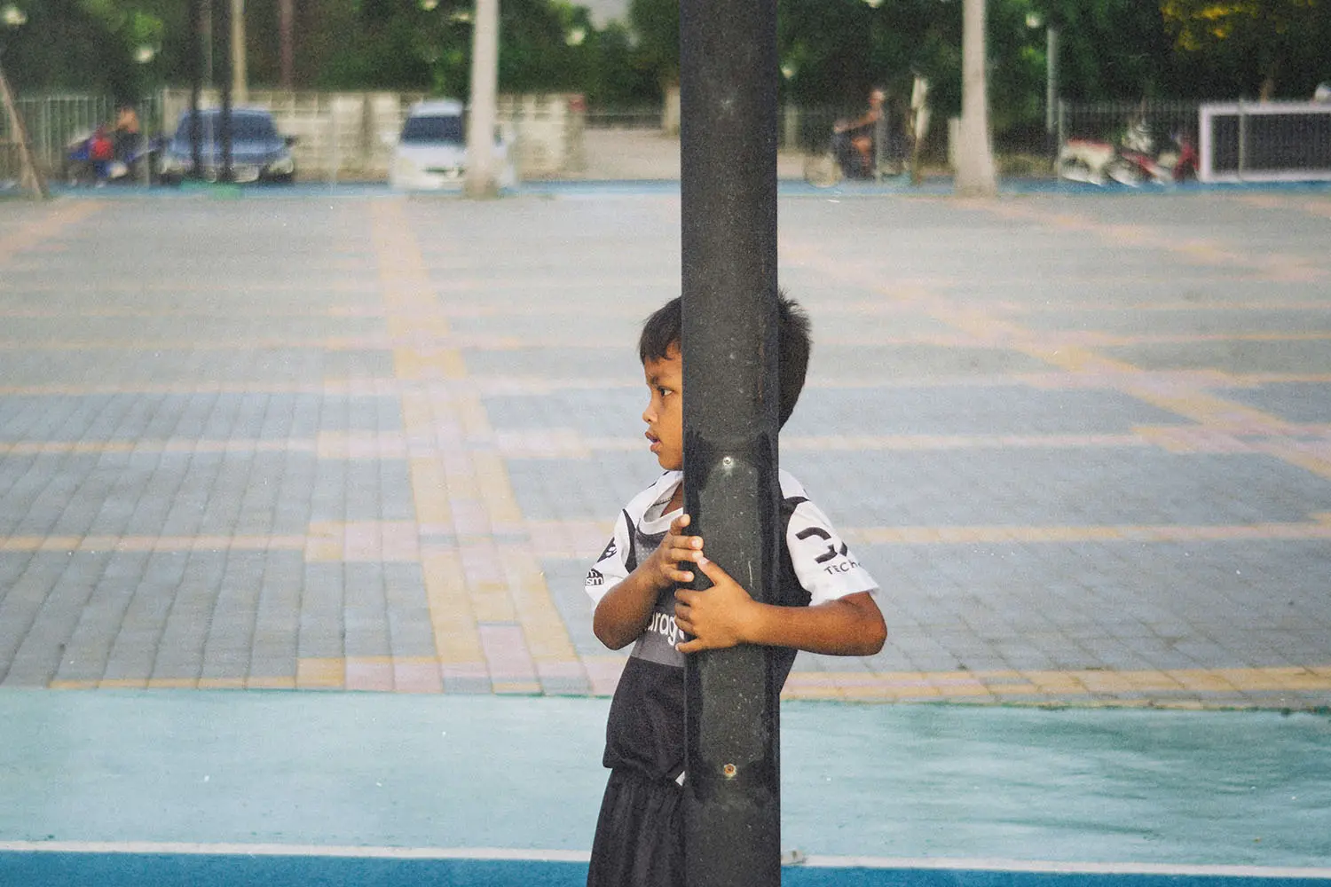 Child wearing a sports uniform stands outdoors, holding onto a tall metal pole in a paved courtyard or playground
