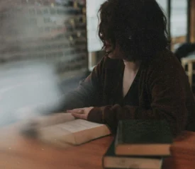 Person sitting at a wooden table reading an open book, with additional books stacked nearby in a softly lit room
