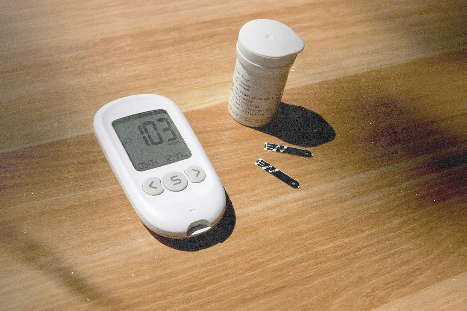 Blood glucose meter reading “103” with test strips and a container on a wooden table