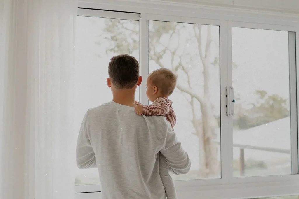 Adult holding a baby while standing by a large window, both looking outside at a softly lit, overcast landscape