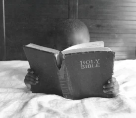 A child sits on a bed holding a large Holy Bible open, with only the book and small hands visible in the foreground