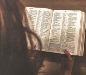 Person reading an open Bible at a wooden table, viewed from overhead with softly lit pages