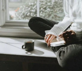Person sits cross‑legged on a windowsill, writing in a notebook beside a mug of coffee in the sunlight