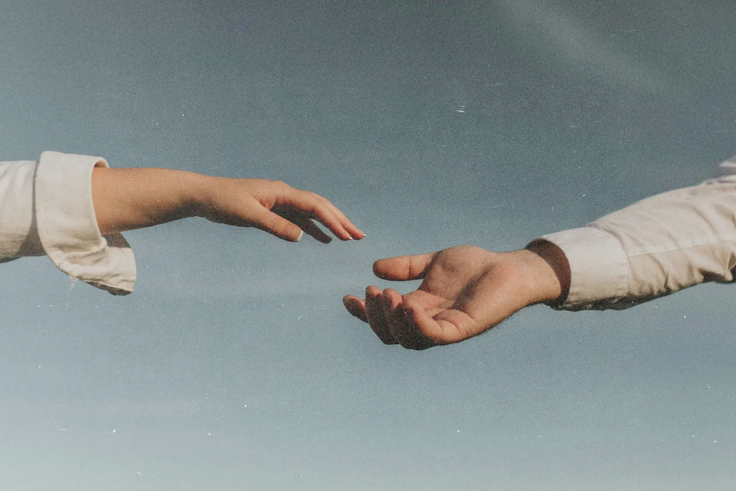 Two outstretched hands reaching toward each other against a soft blue sky, as if about to connect