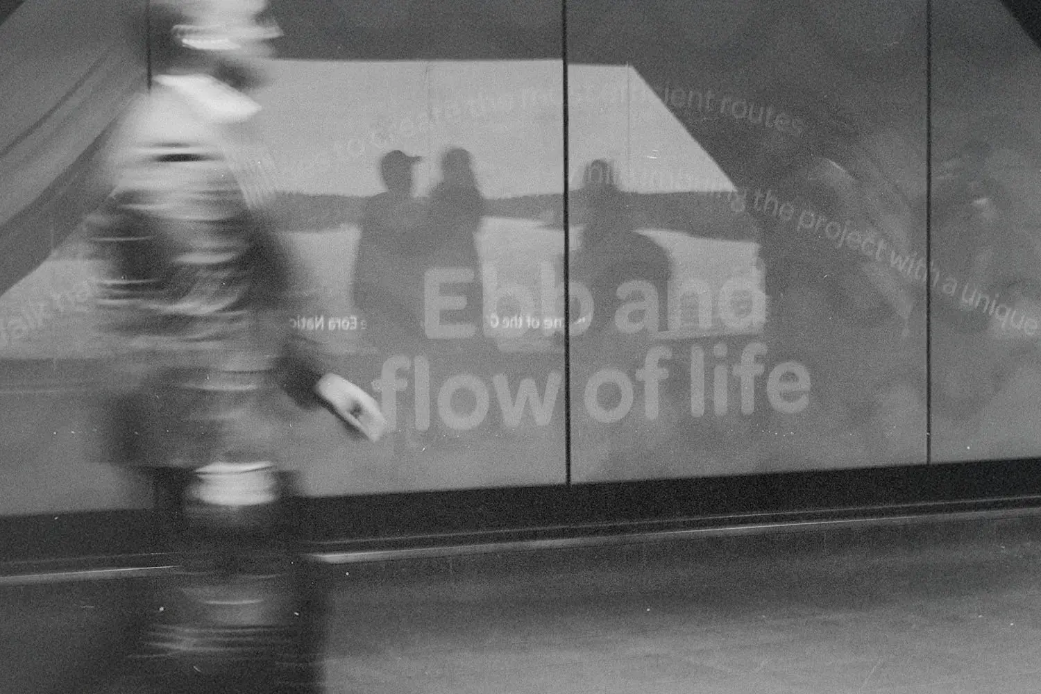 A blurred figure walks past a reflective wall displaying the words “Ebb and flow of life,” with silhouettes visible in the reflection