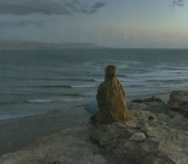 A person sits on a rocky cliff at dusk, watching waves roll across the ocean below