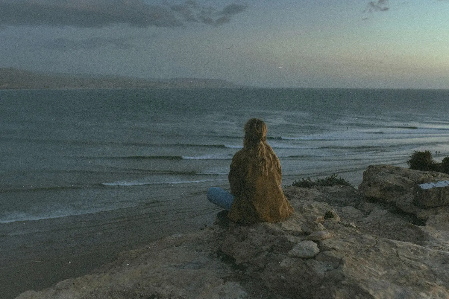 A person sits on a rocky cliff at dusk, watching waves roll across the ocean below