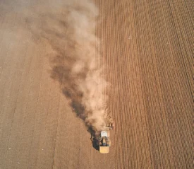 A tractor moves across a large plowed field, leaving a cloud of dust trailing behind it