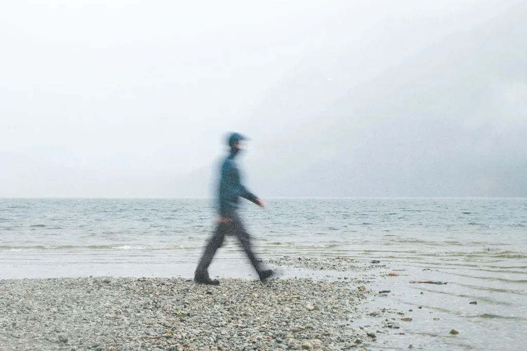 Person walking along a rocky lakeshore on a foggy day with mountains fading into the mist