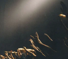 Several raised hands reaching upward toward a bright spotlight in a dark, crowded setting