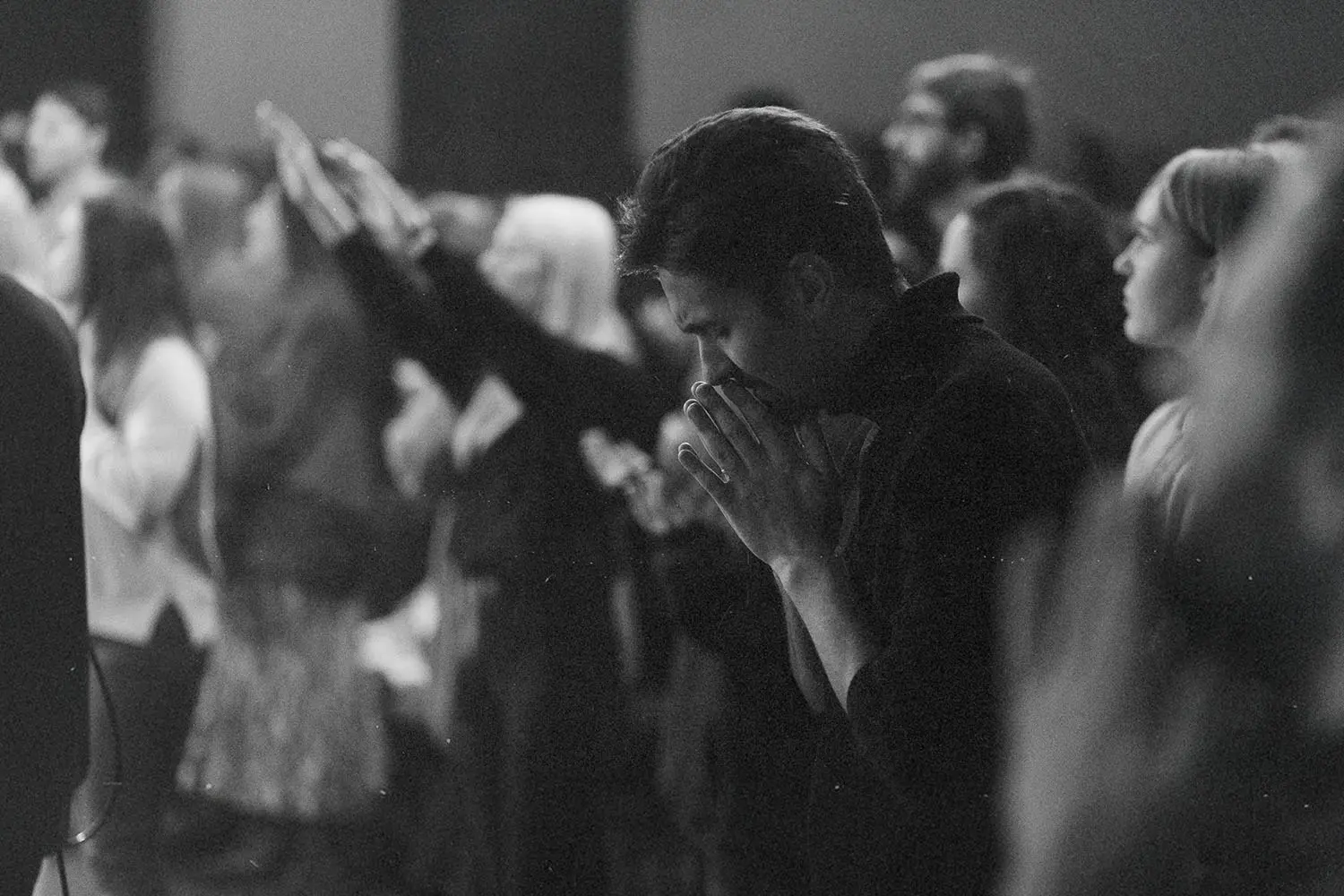 Group of people standing closely together in a dimly lit room with several individuals raising their hands and one person in the foreground holding their hands together in front of their face