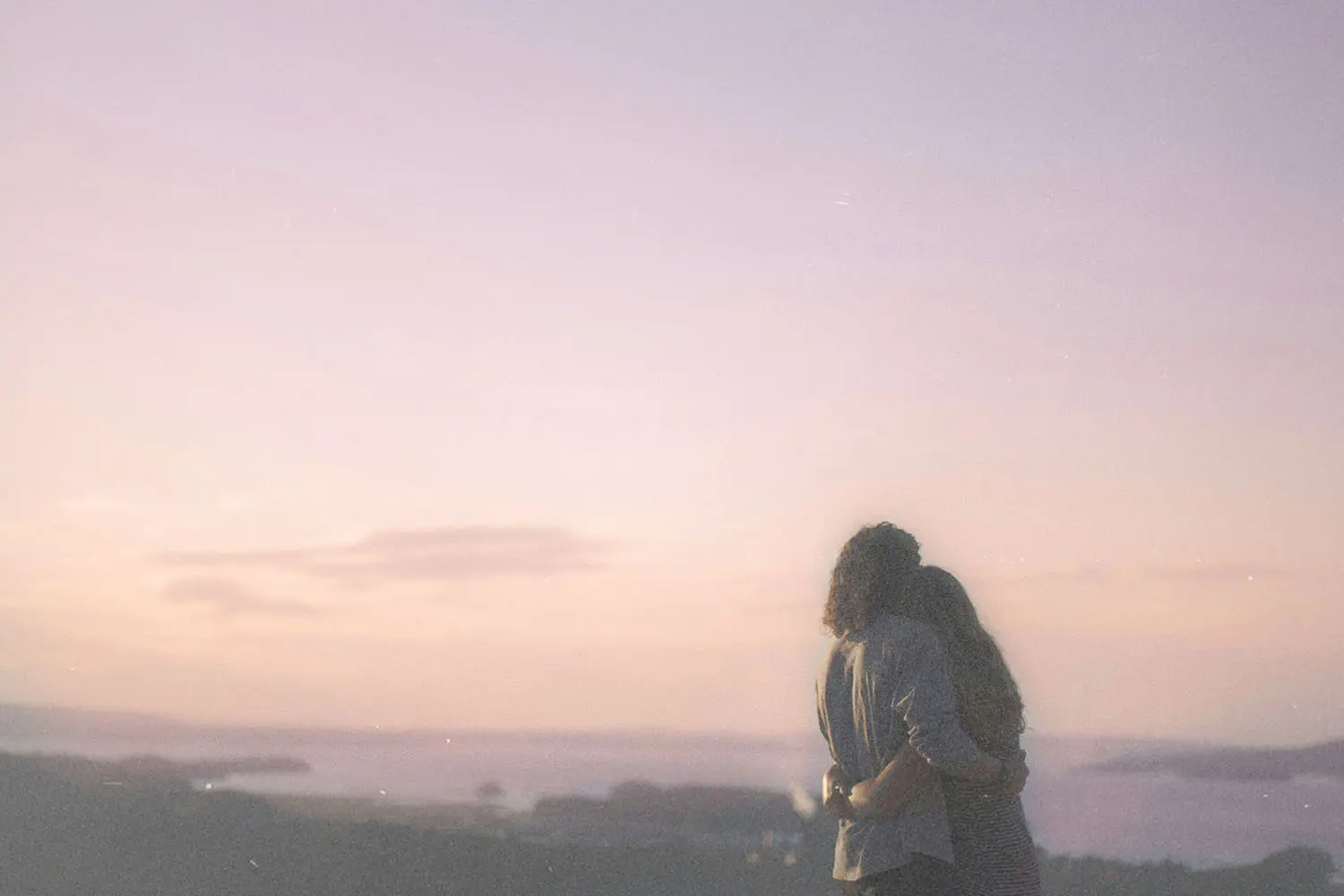Two people standing close together and embracing while looking out over a hazy landscape at sunset, with soft pastel colors in the sky