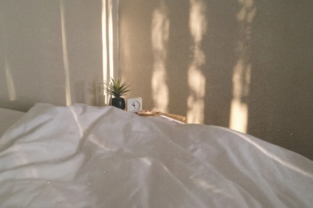 Soft morning light casting vertical shadows on a beige wall behind a bed with rumpled white sheets, a small potted plant, and a clock on the nightstand