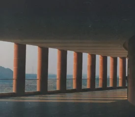 Long covered walkway with tall red columns casting angled shadows across the floor, overlooking a distant landscape in soft morning light