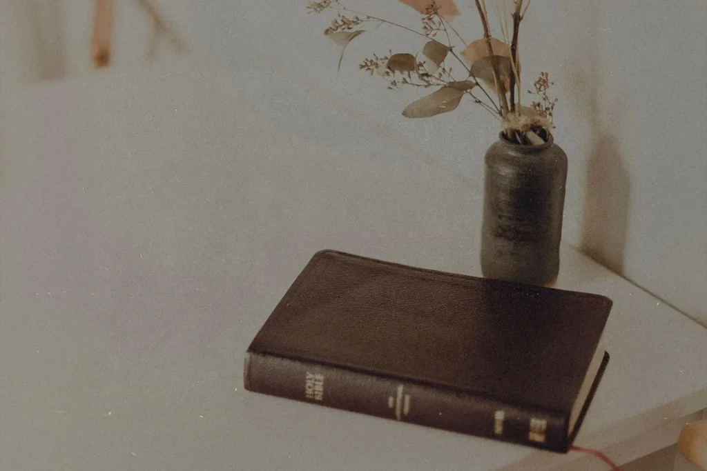Closed Bible rests on a light-colored table beside a small vase filled with dried branches and leaves
