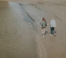 Two people walk side by side along the wet sand near the water’s edge, their reflections visible in the shallow ripples