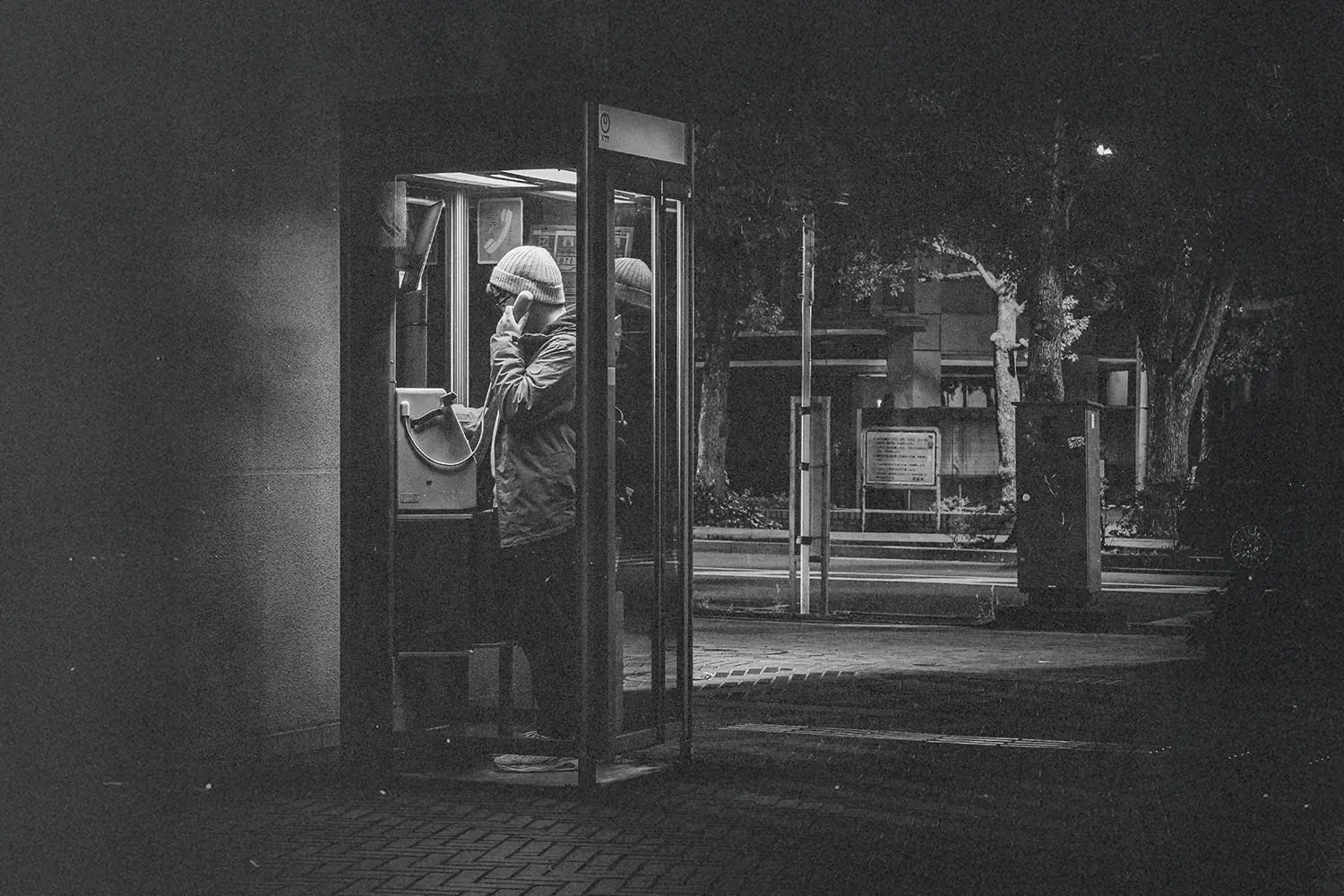 Person standing in a lit phone booth at night on a quiet street