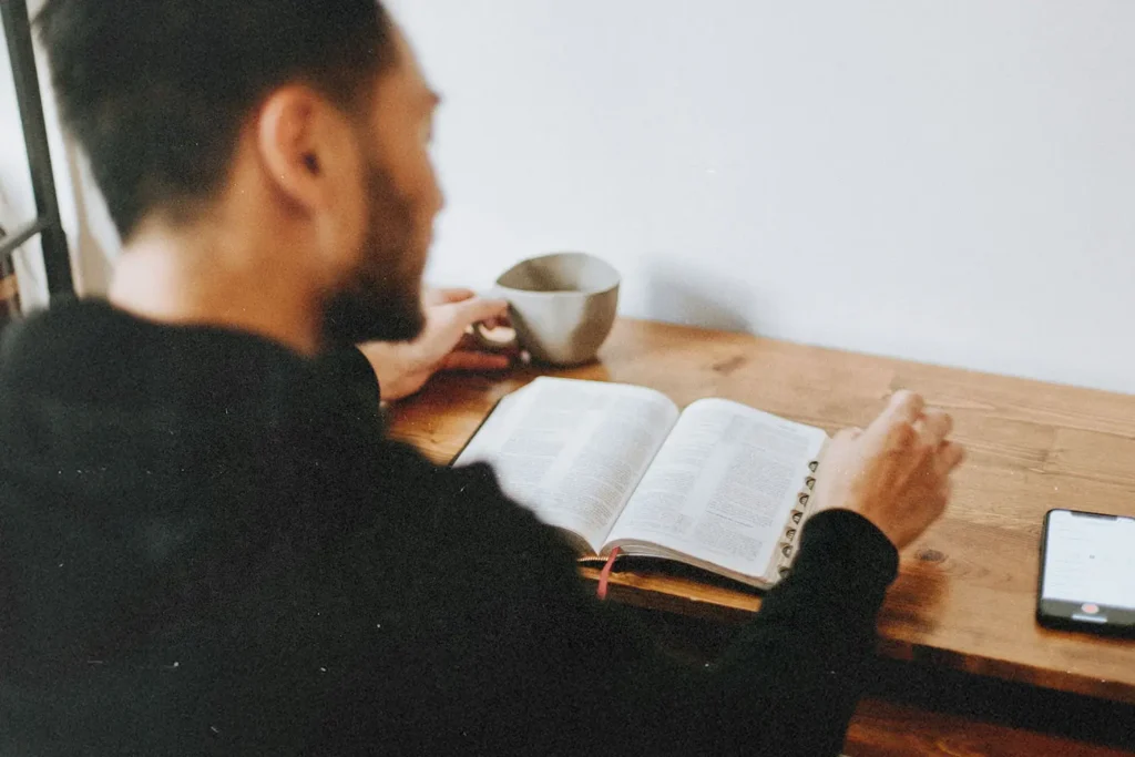 A person sits at a wooden table reading an open Bible with a mug beside them and a smartphone resting nearby
