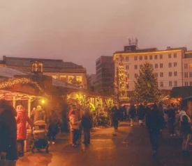 Crowds walking through an outdoor Christmas market at dusk, with warm lights glowing from decorated stalls and buildings in the background