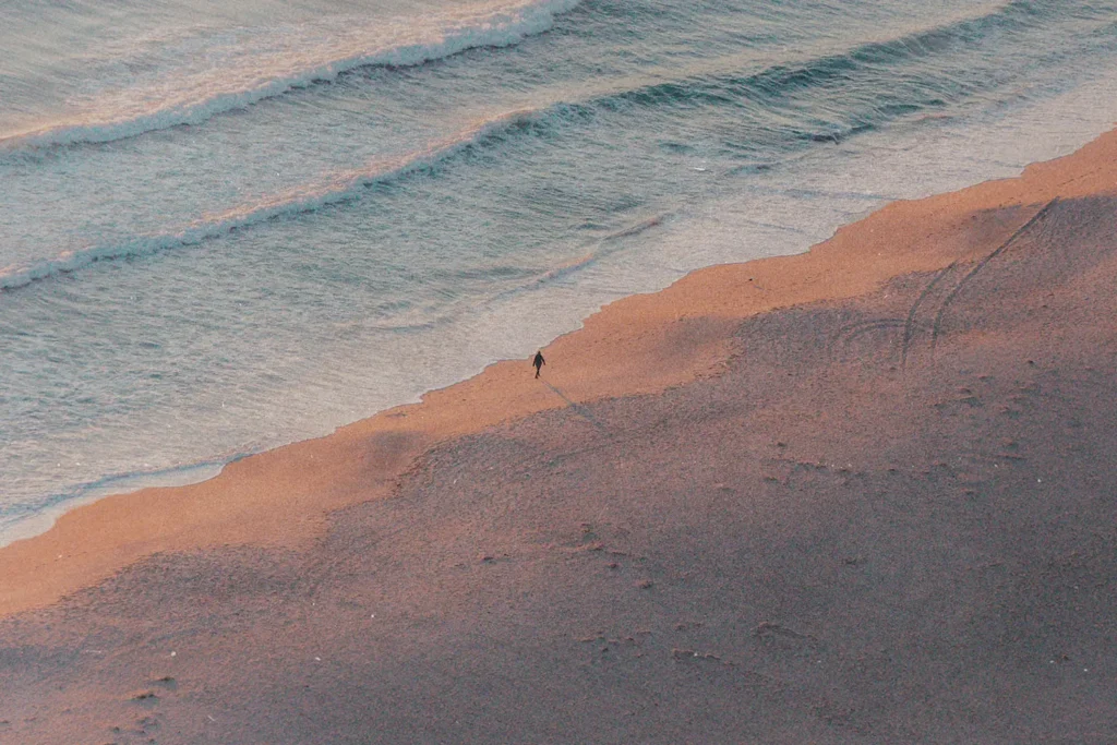 A single person walks along the shoreline at sunrise or sunset, casting a long shadow across the sand as gentle waves roll in