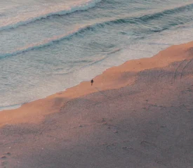 A single person walks along the shoreline at sunrise or sunset, casting a long shadow across the sand as gentle waves roll in