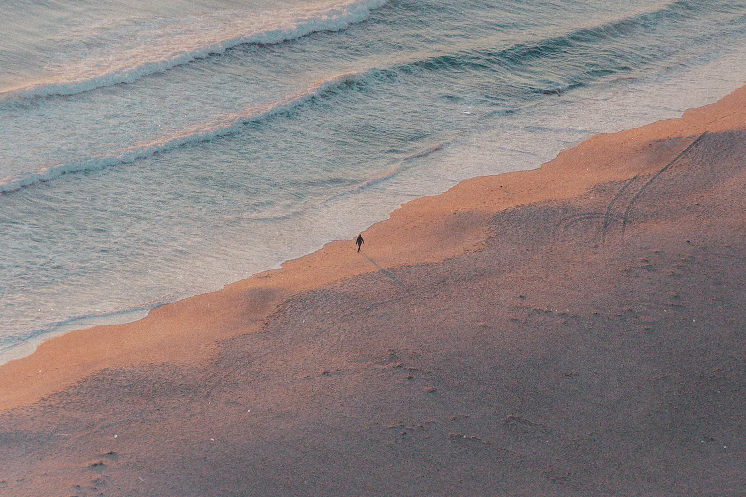 A single person walks along the shoreline at sunrise or sunset, casting a long shadow across the sand as gentle waves roll in
