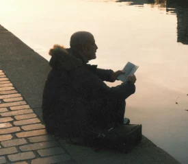 Person sitting on a riverside ledge at sunset, reading a book with calm water reflecting the warm light