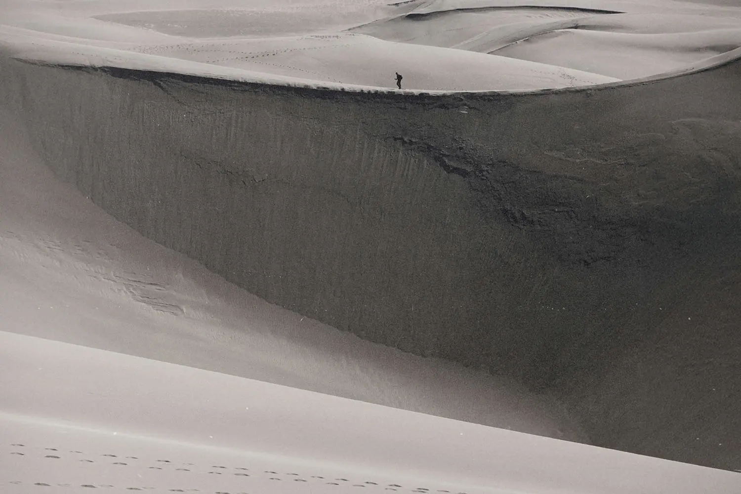 Person walking along the crest of a massive sand dune in a vast, windswept desert landscape