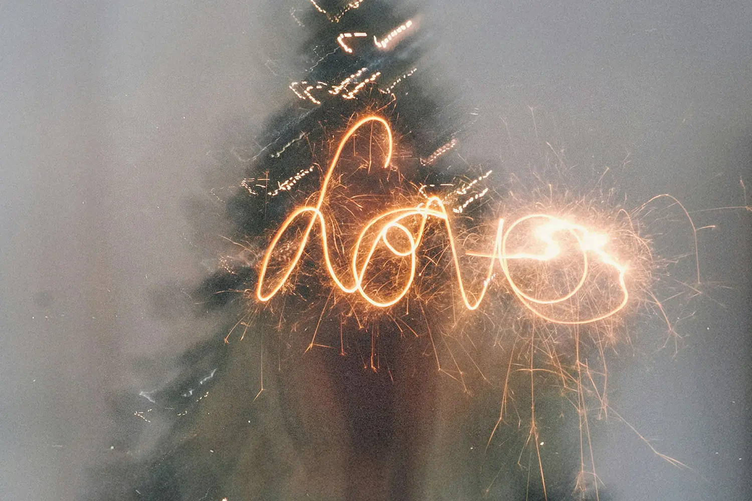 A sparkler writes the word “love” in glowing light trails in front of a blurred Christmas tree