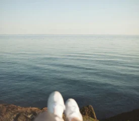 A calm, open body of water stretches to the horizon under soft daylight, with a pair of white shoes visible at the bottom edge of the image