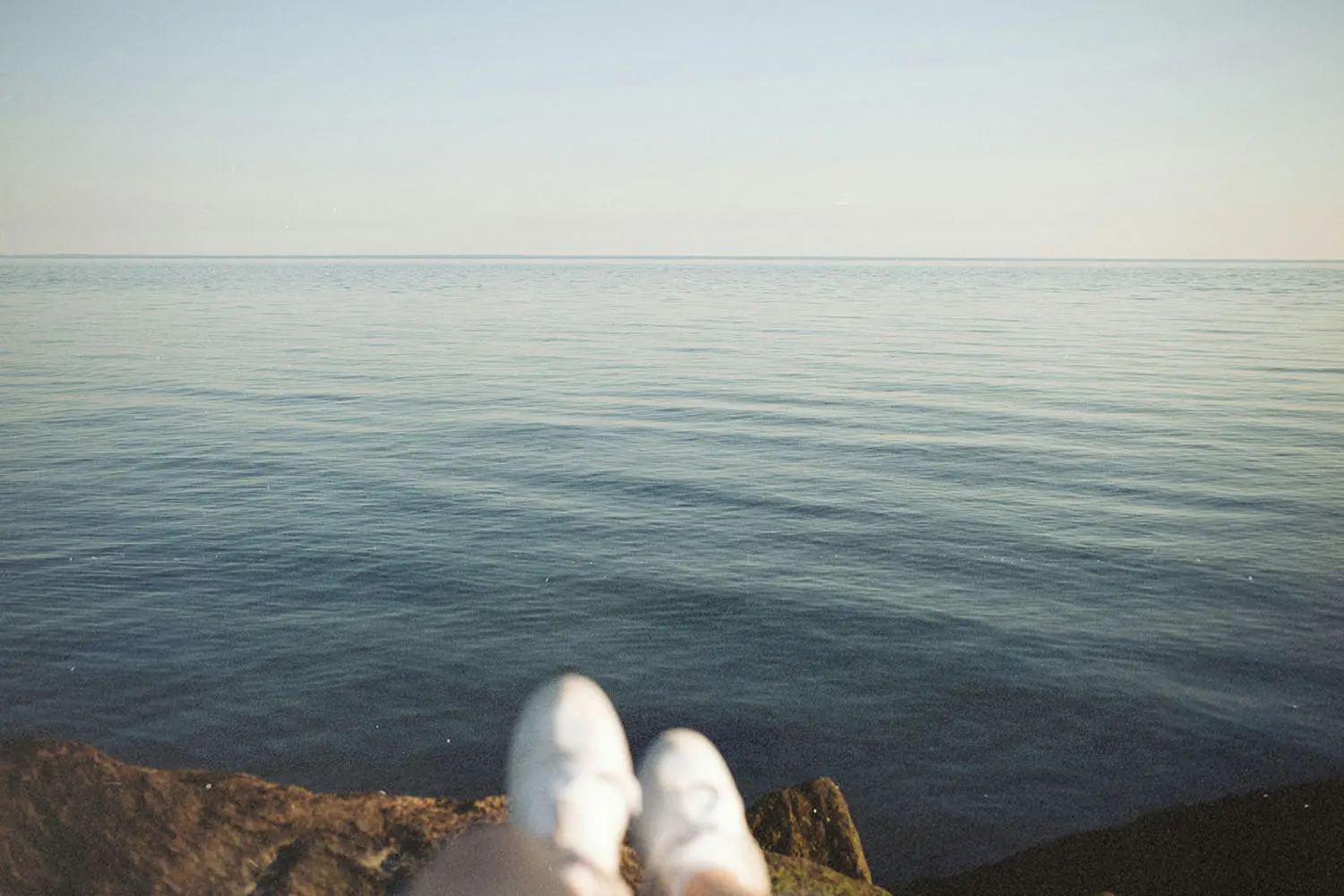 A calm, open body of water stretches to the horizon under soft daylight, with a pair of white shoes visible at the bottom edge of the image