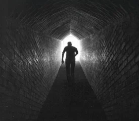 A person walks through a long, dark brick tunnel toward a bright opening at the far end