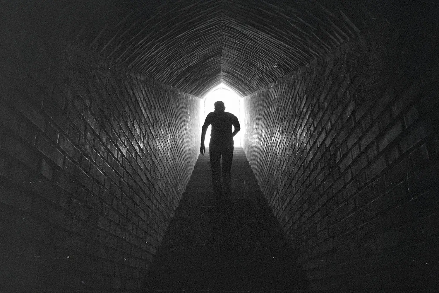 A person walks through a long, dark brick tunnel toward a bright opening at the far end