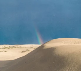 Tall sand dunes under a clear blue sky with a faint rainbow appearing near the horizon