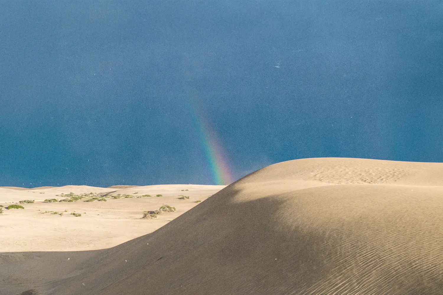 Tall sand dunes under a clear blue sky with a faint rainbow appearing near the horizon