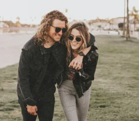 Two people walk arm‑in‑arm across a grassy area near a waterfront path, dressed casually in jackets as they lean affectionately toward each other