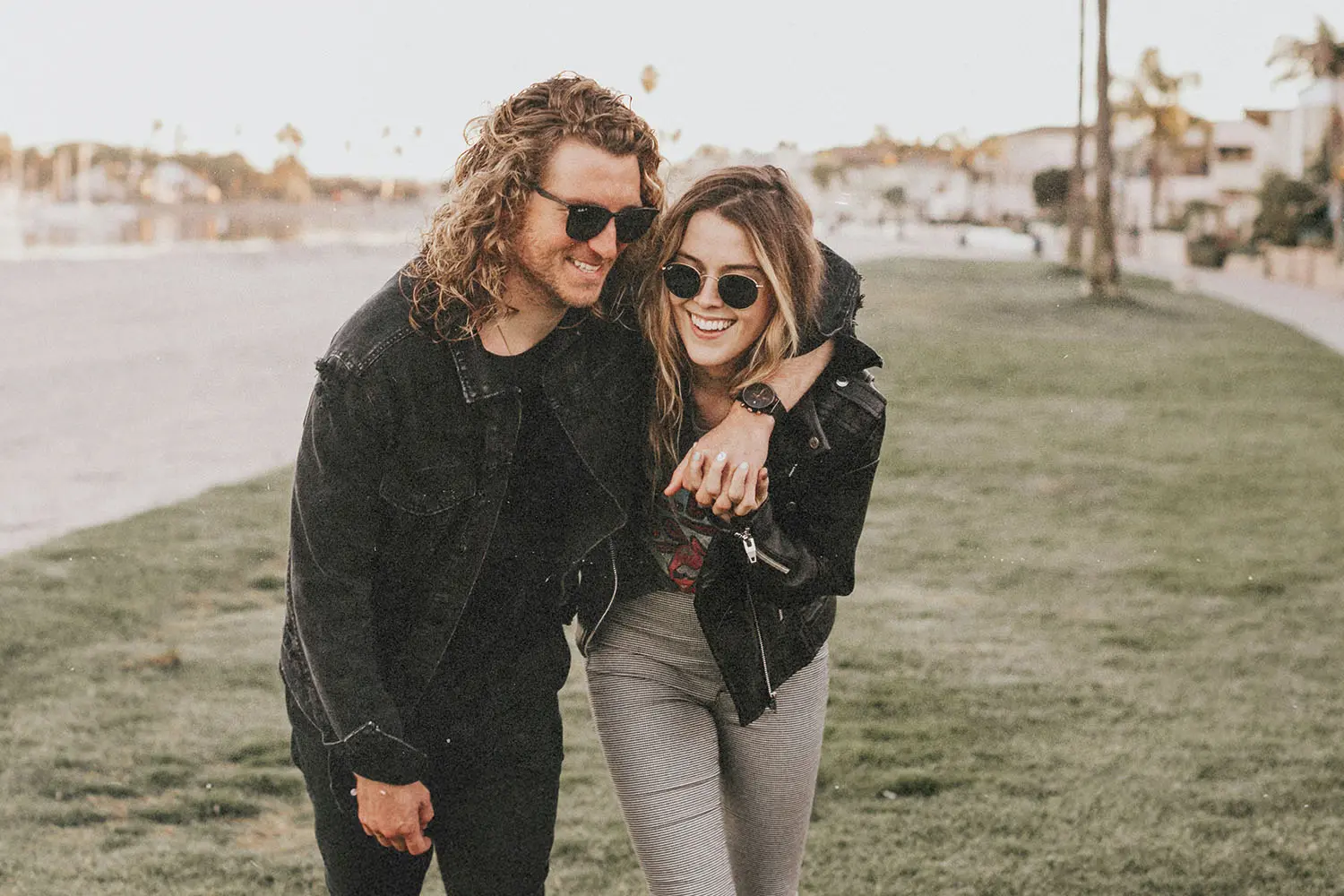 Two people walk arm‑in‑arm across a grassy area near a waterfront path, dressed casually in jackets as they lean affectionately toward each other