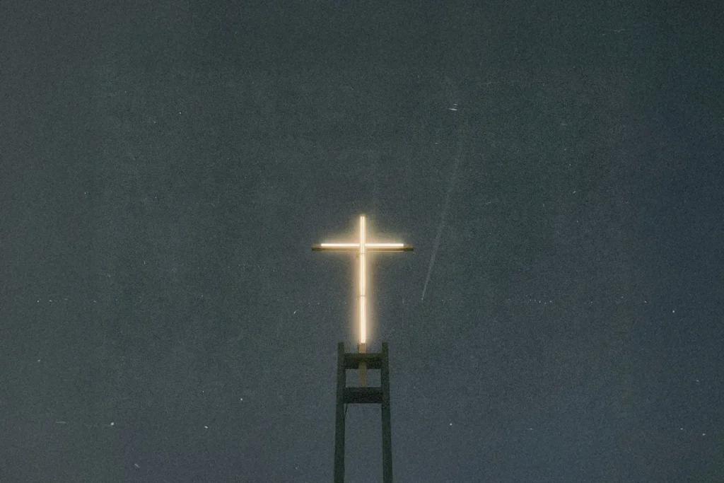 A glowing cross sits atop a tall structure against a dark, starless night sky